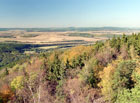 V dálce je vidět zelená plocha chráněné krajinné oblasti Křivoklátsko s vyčnívajícími kopci Velíz a Krušná hora.

