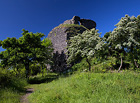 Pohled směrem k Hazmburku. Ruiny středověkého hradu Košťálov trůní na vysoké čedičové skále neobvykle příkře se zvedající nad své okolí. Výrazný skalnatý vrchol doslova vyčnívá mezi sousedními zaoblenějšími kupovitými kopci Českého středohoří.

